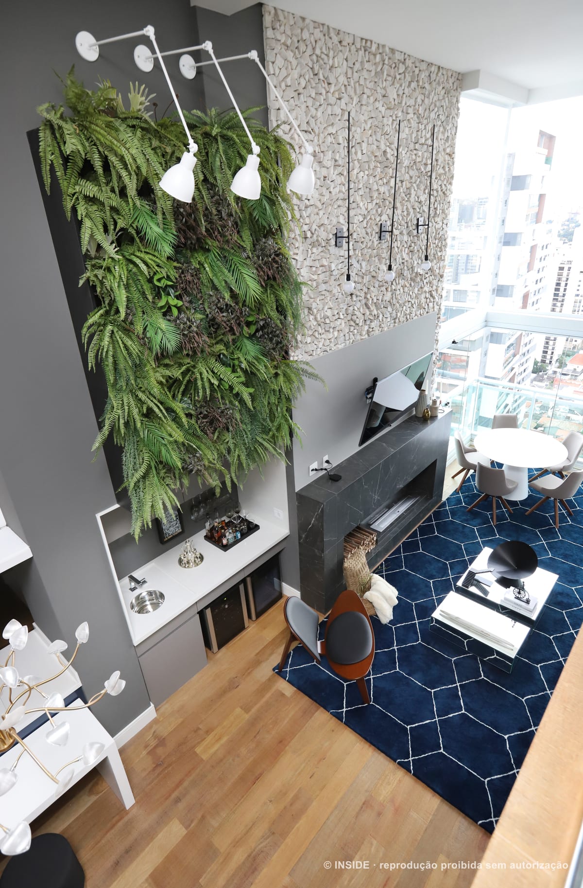 Parede de vegetação em pé-direito duplo sobre o estar, tapete azul geométrico, lareira de pedra e a vista do bairro pela vidraça, apartamento Brooklin-PE, foto Mariana Orsi, acervo INSIDE