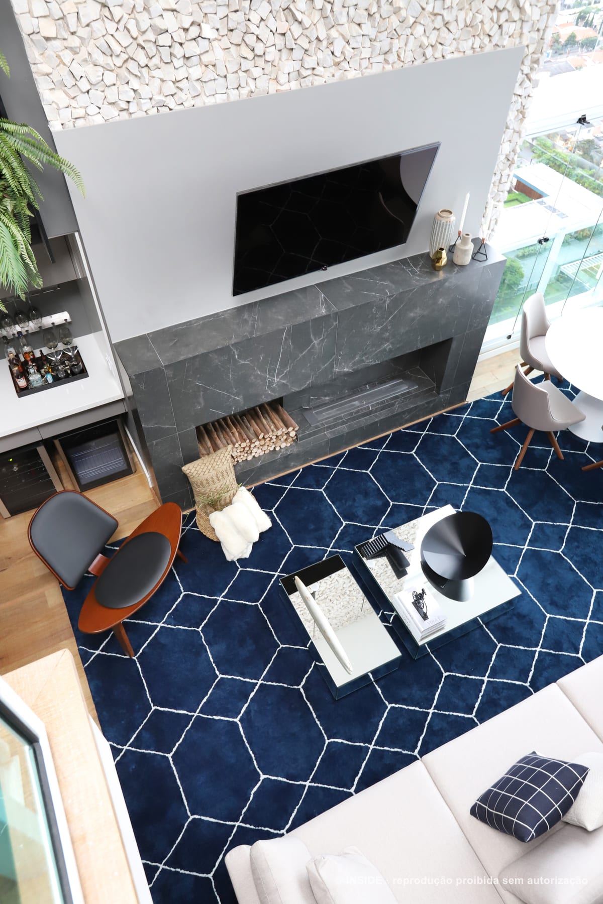Estar com lareira revestida em porcelanato que imita mármore, poltrona laranja de encosto curvo, tapete azul de losangos e a cidade pela vidraça, apartamento Brooklin-PE, foto Mariana Orsi, acervo INSIDE
