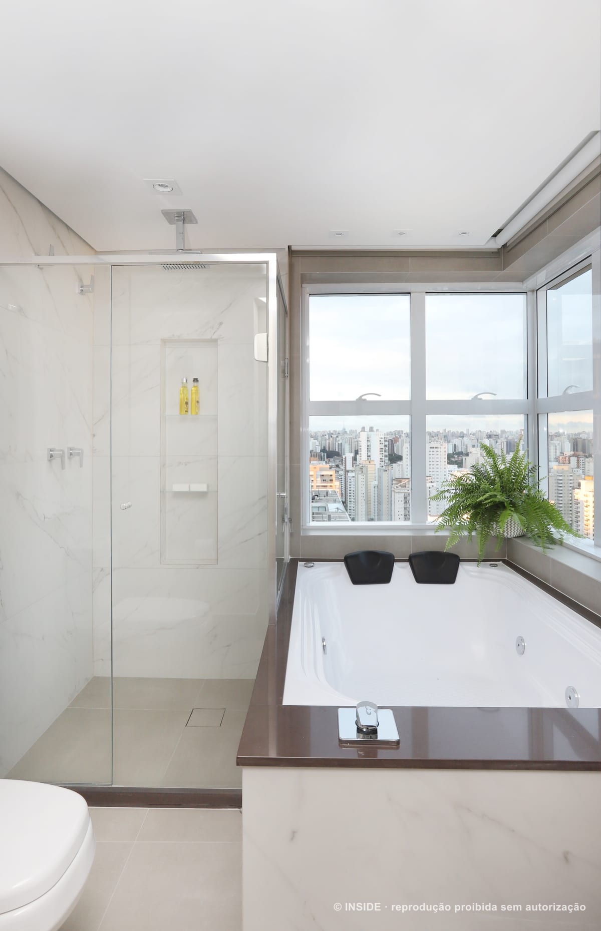 Banheiro com banheira junto à janela de canto, revestimento branco marmorizado e um vaso de samambaia no parapeito, com a cidade ao fundo, apartamento Brooklin-PE, foto Mariana Orsi, acervo INSIDE