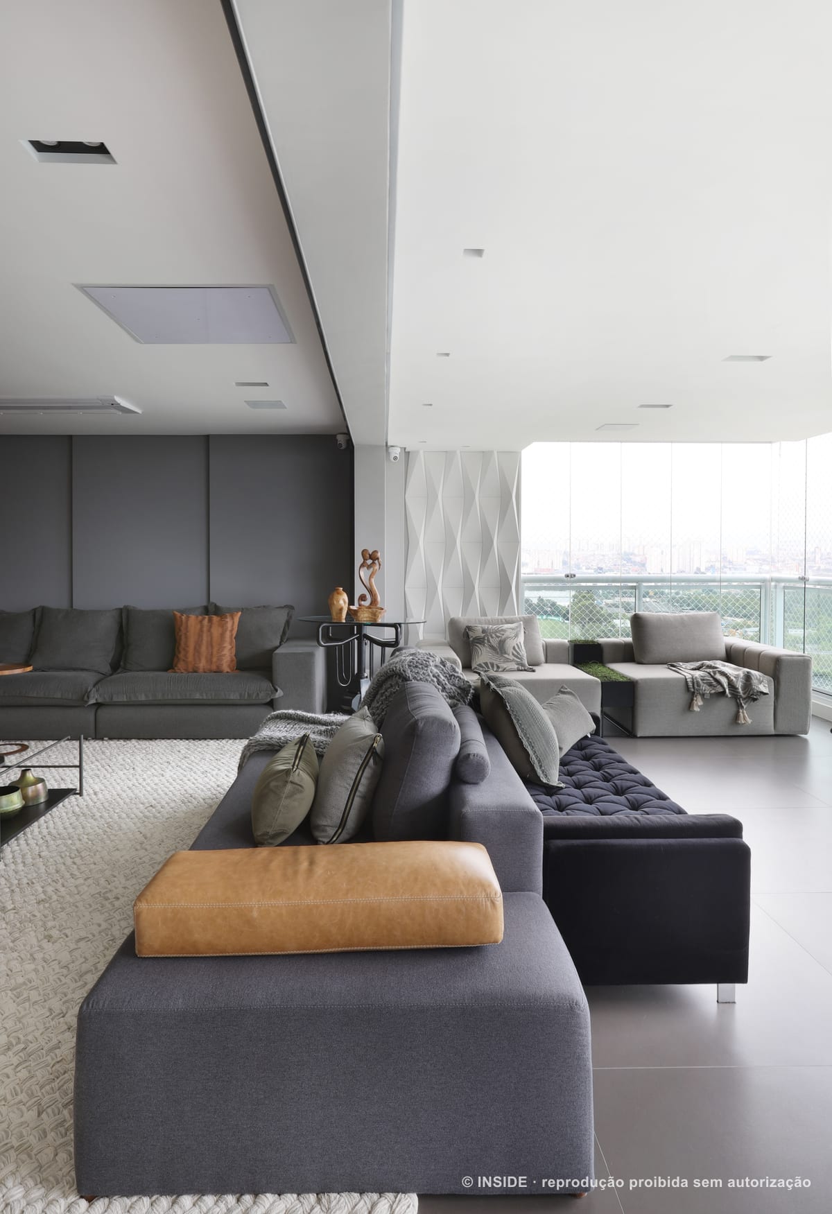 Sala de estar ampla e integrada, com sofá cinza em L, chaise de couro caramelo, painel geométrico branco e a cidade pela vidraça, apartamento Perdizes-DB, foto Mariana Orsi, acervo INSIDE