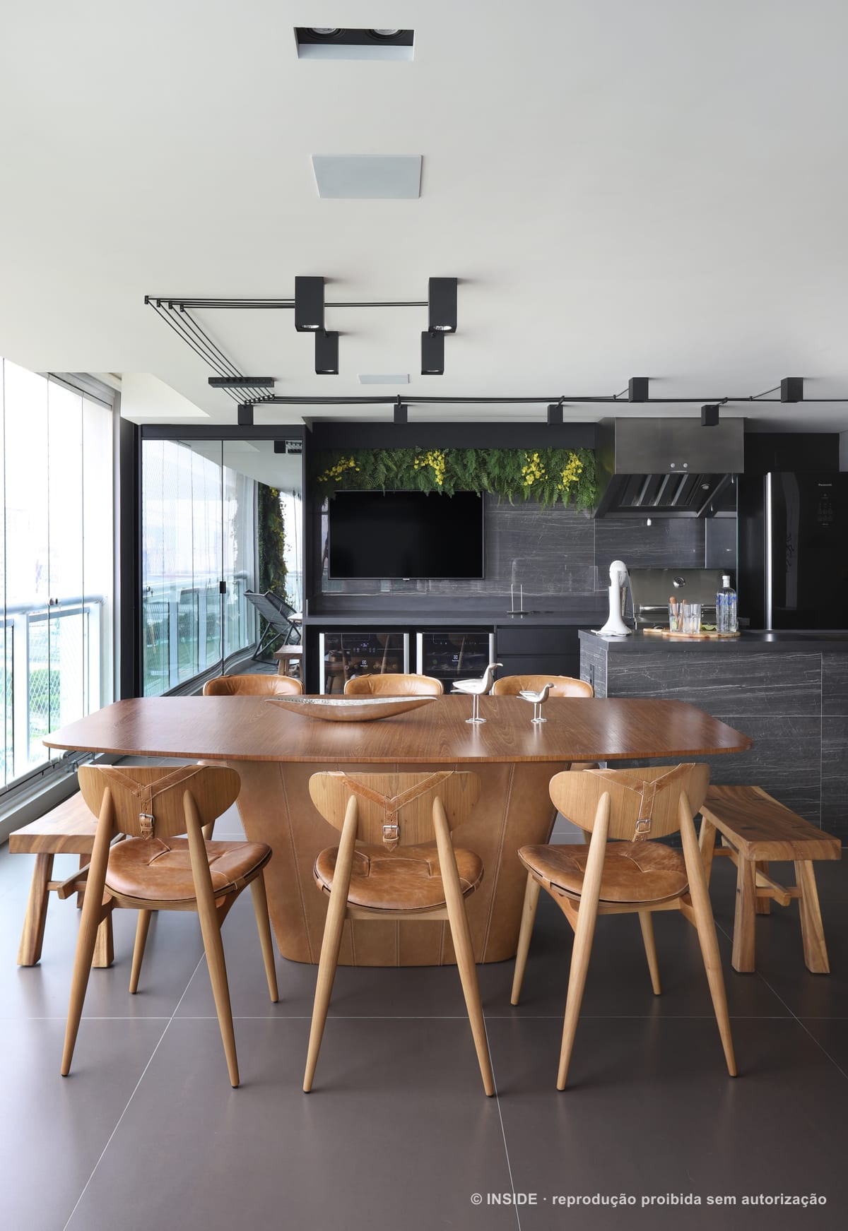 Mesa oval de madeira com cadeiras Wishbone na varanda envidraçada, sob jardim vertical e televisão, apartamento Perdizes-DB, foto Mariana Orsi, acervo INSIDE