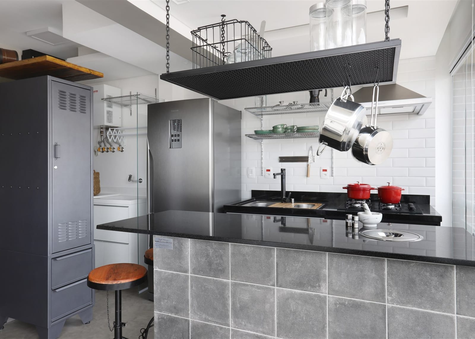 Cozinha aberta com ilha revestida de cimento, bancada de granito preto, cooktop a gás, cuba embutida e estrutura suspensa de panelas ancorada no teto, projeto INSIDE