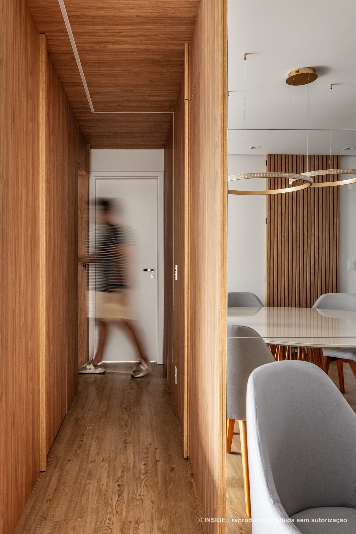 Corredor de madeira do apartamento Amaro-BJ, com alguém atravessando em movimento e, ao lado, a mesa de jantar sob o pendente de argolas, acervo INSIDE