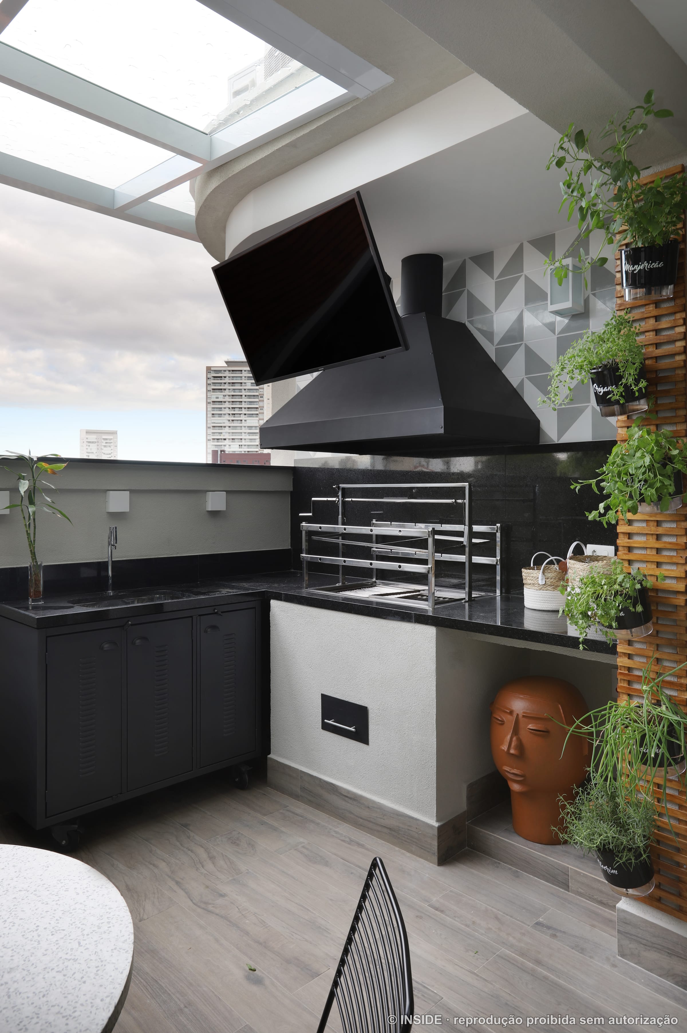Varanda gourmet com churrasqueira de granito preto, coifa preta, painel treliçado com vasos de ervas e vista da cidade, projeto INSIDE