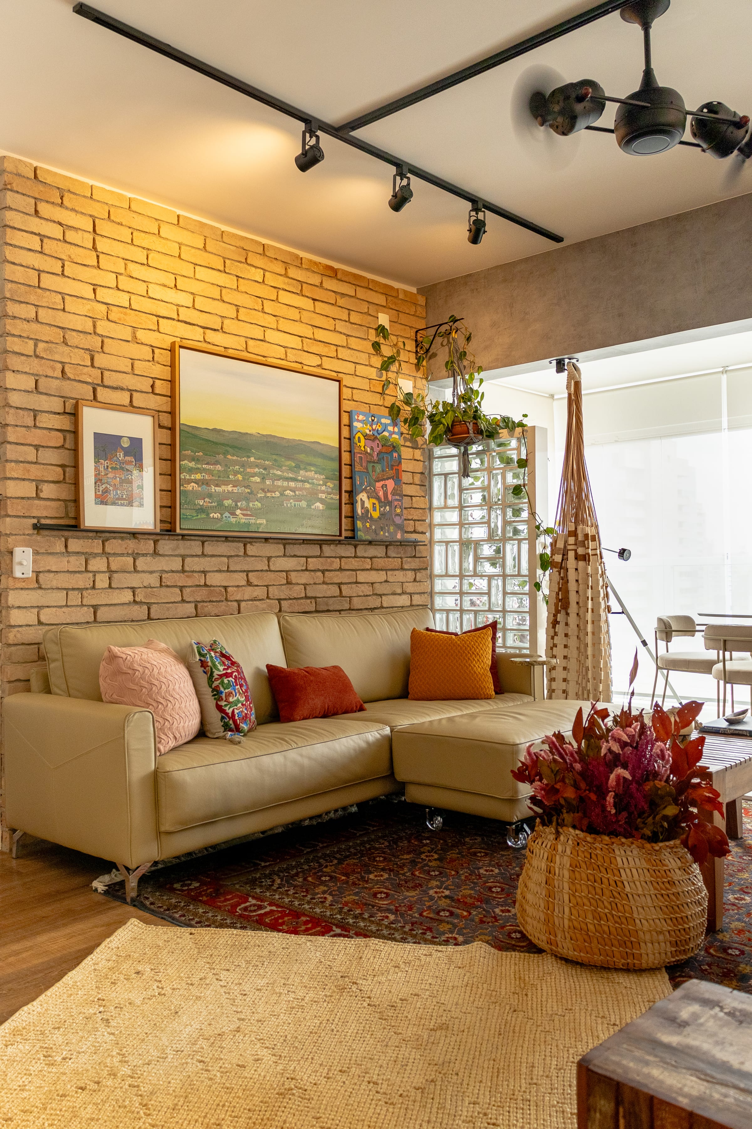 Sala boêmia com parede de tijolo aparente, sofá de couro caramelo em L, trio de quadros naïf, tapete persa e rede de macramê, projeto INSIDE