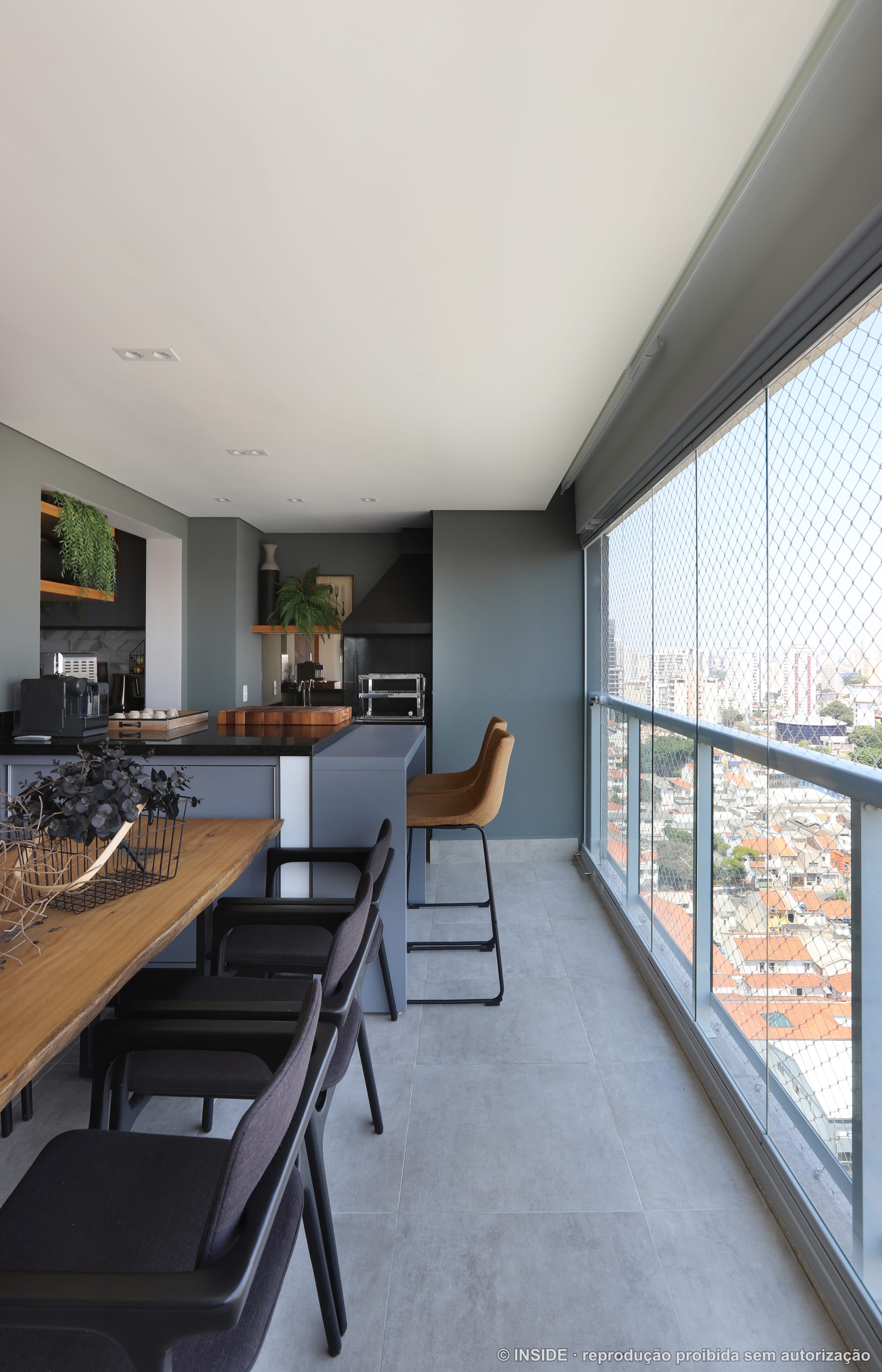 Varanda gourmet com paredes verde escuro, bancada de granito preto, mesa de madeira maciça e vista para a cidade, projeto INSIDE