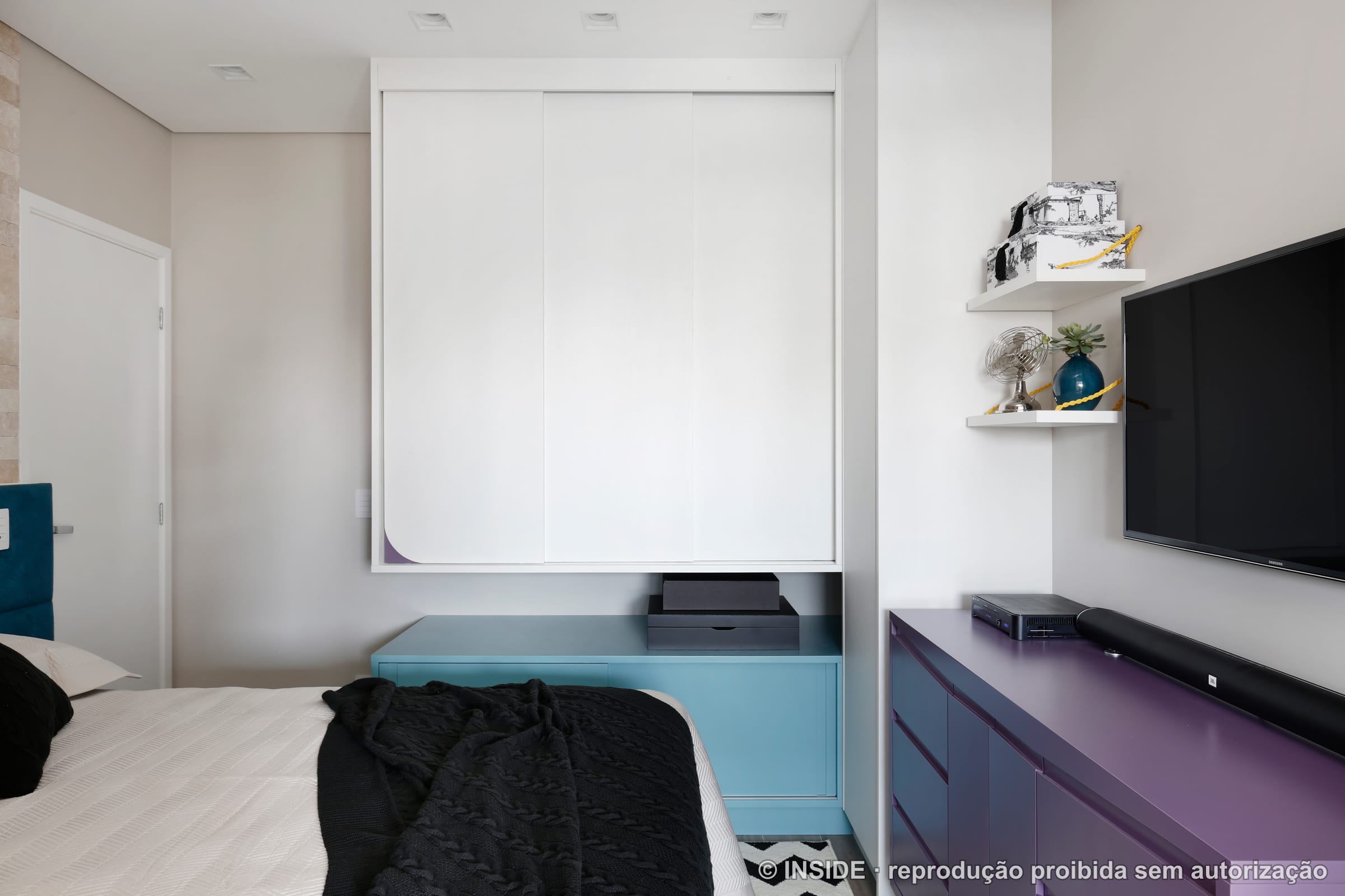 Suíte vista do lado do armário embutido branco, com rack roxo, TV e balcão azul claro, projeto INSIDE